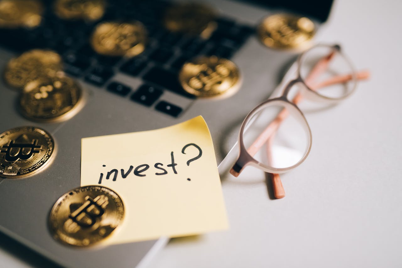 hero-img-01 A close-up of bitcoin coins on a laptop with eyeglasses and a sticky note reading 'invest?'